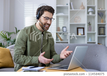 Man with a headset conducting a virtual business meeting from home, sitting on a couch with a laptop and notebook. Man with a headset conducting a virtual business meeting from home, sitting on a couch with a laptop and notebook. 114584075
