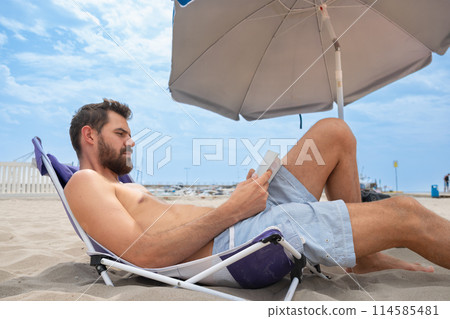 Young adult man with beard reading focused lying on beach with umbrella Young adult man with beard reading focused lying on beach with umbrella 114585481