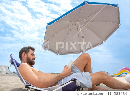 Young man reading focused lying on beach with umbrella and inflatable float 114585484
