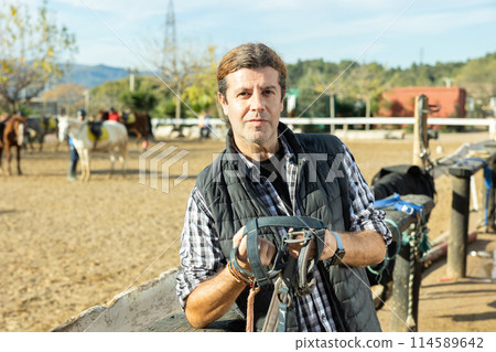 Middle-aged man instructor holding horse halter or reins during working day at horse club on sunny day of spring Middle-aged man instructor holding horse halter or reins during working day at horse club on sunny day of spring 114589642