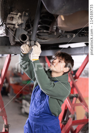 Young mechanic repairing undercarriage of vehicle on lift in auto repair workshop 114589735