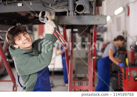 Smiling young mechanic repairing chassis of car on lift in workshop Smiling young mechanic repairing chassis of car on lift in workshop 114589788
