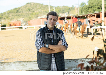Portrait of confident horseman in plaid shirt posing on yard of horse riding club on sunny day of autumn 114590167
