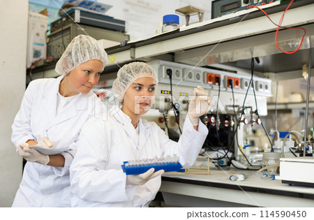 Adult and young female chemists examine liquid in beaker in laboratory Adult and young female chemists examine liquid in beaker in laboratory 114590450