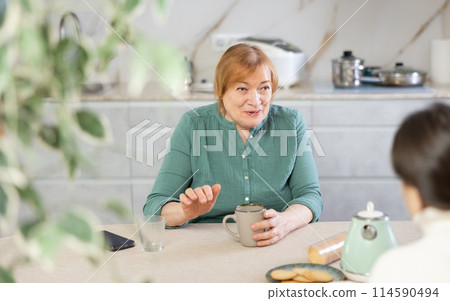 In kitchen at table, elderly smiling woman communicates with friend and drinks coffee or tea In kitchen at table, elderly smiling woman communicates with friend and drinks coffee or tea 114590494
