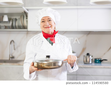 Elderly woman in chef hat and white cook uniform stands near kitchen table and holds frying pan Elderly woman in chef hat and white cook uniform stands near kitchen table and holds frying pan 114590580