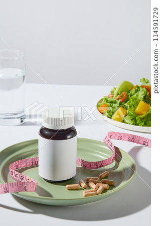 A medicine bottle without label displayed on a round green plate with some yellow pills and a pink measuring tape, a dish of vegetable salad and a glass of water placed in back on white background 114591729