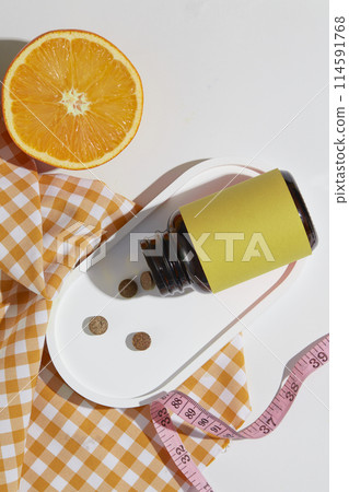 Top view photography at a white flat contains some preparation items for weight loss therapy, medicine bottle with yellow blank label, yellow checkered towel and orange slice. Vacant space for design 114591768