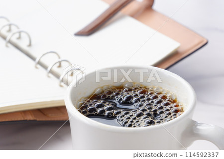 照片中，一個人喝著現煮的咖啡，打開環形筆記本，思考自己的工作行程。 114592337