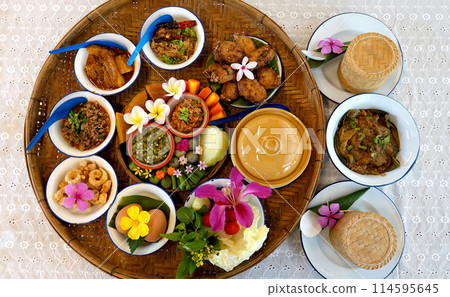 Northern Thai cuisine food set. Pork curry, chilli dip, minced pork salad, soft boiled vegetables and pork snack served in brass dishes. Decorated with flower, garland, spoon and basket background. 114595645