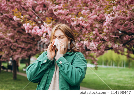 Sneezing young redhead woman with nose wiper among blooming trees in park. Portrait of sick women sneezes in white tissue, suffers from rhinitis and running nose. Symptoms of cold or allergy. Sneezing young redhead woman with nose wiper among blooming trees in park. Portrait of sick women sneezes in white tissue, suffers from rhinitis and running nose. Symptoms of cold or allergy. 114598447