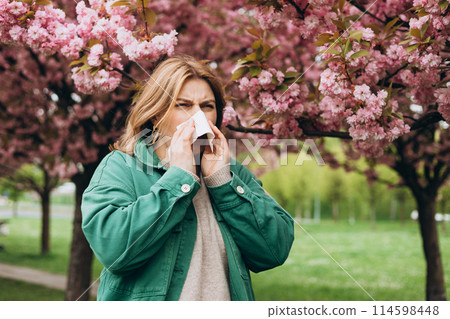 Sneezing young redhead woman with nose wiper among blooming trees in park. Portrait of sick women sneezes in white tissue, suffers from rhinitis and running nose. Symptoms of cold or allergy. Sneezing young redhead woman with nose wiper among blooming trees in park. Portrait of sick women sneezes in white tissue, suffers from rhinitis and running nose. Symptoms of cold or allergy. 114598448