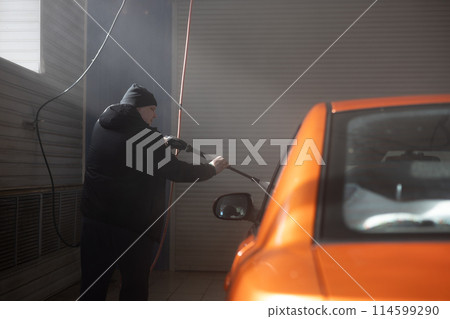 At a car wash, a man washes off dirt and dust from his car. A man walks around a car and washes an orange car with water under pressure. At a car wash, a man washes off dirt and dust from his car. A man walks around a car and washes an orange car with water under pressure. 114599290