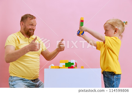 Little son showing with his father his little building while they playing against pink studio background. Man showing thumb up. 114599806