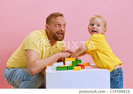 Father guides his little boy, son in building toy with vivid blocks against pink studio background. 114599816