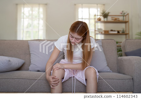 Young Asian woman experiencing knee pain while sitting on a sofa at home. Concept of health issues and discomfort 114602343