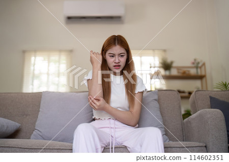Young Asian woman experiencing arm pain while sitting on a sofa at home. Concept of health issues and discomfort Young Asian woman experiencing arm pain while sitting on a sofa at home. Concept of health issues and discomfort 114602351
