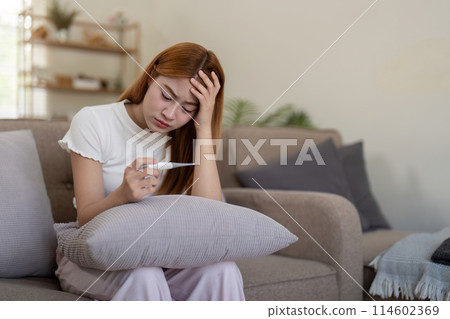 Young Asian woman with fever checking her temperature with a thermometer at home. Concept of illness and healthcare Young Asian woman with fever checking her temperature with a thermometer at home. Concept of illness and healthcare 114602369