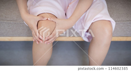Close up of young Asian woman holding her knee in pain while sitting on a sofa. Concept of knee pain and health issues Close up of young Asian woman holding her knee in pain while sitting on a sofa. Concept of knee pain and health issues 114602385