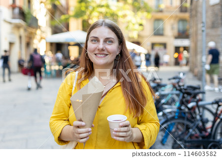 Carefree woman eating traditional Churros, a fried pastry with chocolate on a city street. Positive cheerful female student posing outdoors. Food concept 114603582