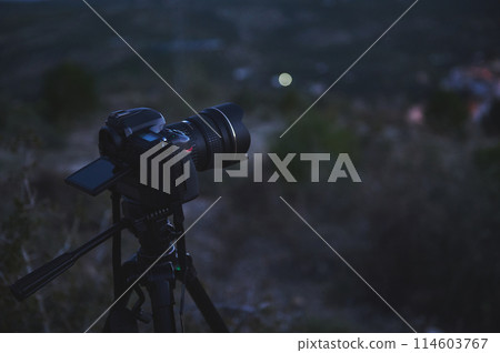A black digital semi-mirror camera photographic equipment, with movable mockup touch screen, placed on tripod in nature, capturing steady picture or recording video of the city in the night and dawn. A black digital semi-mirror camera photographic equipment, with movable mockup touch screen, placed on tripod in nature, capturing steady picture or recording video of the city in the night and dawn. 114603767
