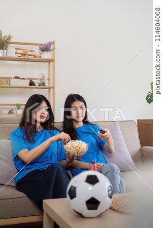 Friends watching a football game at home. Two women enjoying a sports match on TV with popcorn and snacks Friends watching a football game at home. Two women enjoying a sports match on TV with popcorn and snacks 114604008