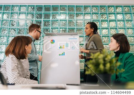 Low-angle view of two skilled multiethnic business speakers give chart presentation at conference meeting in office. Diverse male and female presenters coaches work in teamwork training staff group. 114604499