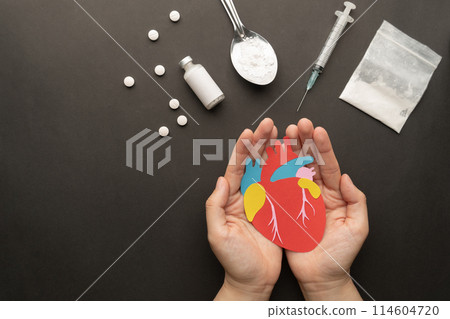 Effects of drugs on the human heart. Hands holding paper cut of heart with drug powder, syringe and pills on dark background. International Day against Drug Abuse. Effects of drugs on the human heart. Hands holding paper cut of heart with drug powder, syringe and pills on dark background. International Day against Drug Abuse. 114604720