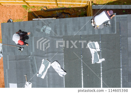 An experienced roofing contractor uses an air nail gun to install new asphalt bitumen shingles on roof of house 114605398