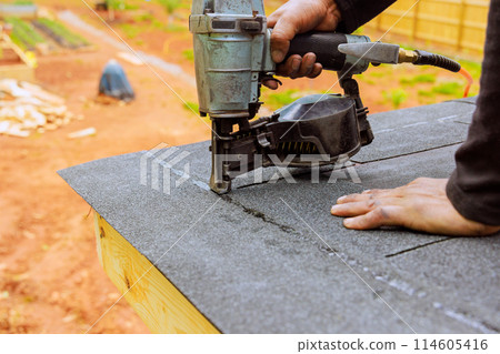 Roofer using an air pneumatic nail gun installs new asphalt bitumen shingles Roofer using an air pneumatic nail gun installs new asphalt bitumen shingles 114605416