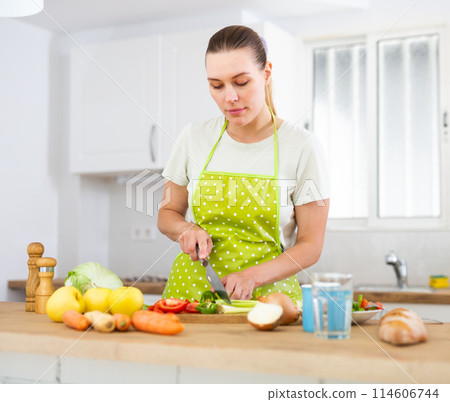 Positive young girl slicing vegetables for salad in cozy kitchen Positive young girl slicing vegetables for salad in cozy kitchen 114606744