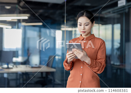 Serious businesswoman thinking and working on a digital tablet in a modern office. Focused and determined Asian woman looking at screen. Serious businesswoman thinking and working on a digital tablet in a modern office. Focused and determined Asian woman looking at screen. 114608381