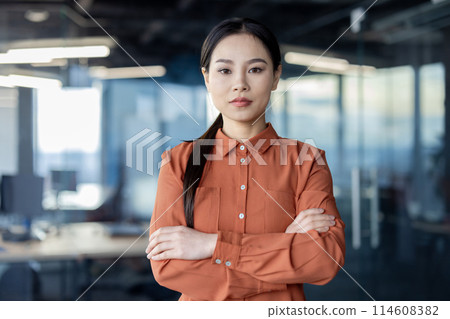 A serious thinking Asian businesswoman in a modern office. She conveys professionalism and confidence with arms crossed, reflecting the work environment. A serious thinking Asian businesswoman in a modern office. She conveys professionalism and confidence with arms crossed, reflecting the work environment. 114608382