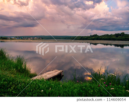 Beautiful pink sunset over calm lake at countryside evening. Tranquil lake with a wooden dock in the foreground. Beautiful pink sunset over calm lake at countryside evening. Tranquil lake with a wooden dock in the foreground. 114609054
