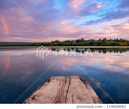 Beautiful pink sunset with rainbow reflection over calm lake at countryside evening. Tranquil lake with a wooden dock in the foreground. 114609063
