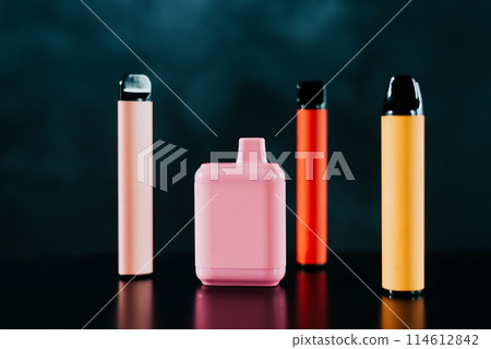 A set of colorful disposable electronic cigarettes of different shapes on a black background with smoke. Concept of modern smoking, vaping and nicotine. 114612842