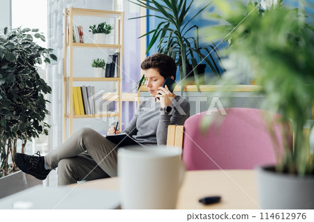 Confident Neutral gender middle aged woman in casual clothing having conversation by phone, making note in notebook in office, meeting room. Phone interview. HR specialist, hiring, recruiting 114612976