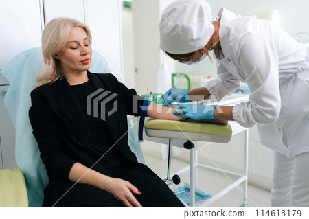 Female nurse sticking needle into female arm for blood sampling. Skilled lab assistant in blue medical gloves inserting needle into young patient arm. Nurse taking venous blood for test in clinic. Female nurse sticking needle into female arm for blood sampling. Skilled lab assistant in blue medical gloves inserting needle into young patient arm. Nurse taking venous blood for test in clinic. 114613179