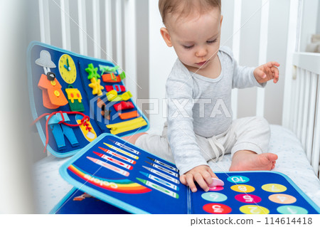 Little toddler girl playing with busy book while sitting in crib. Concept of quiet books and modern educational toys for kids Little toddler girl playing with busy book while sitting in crib. Concept of quiet books and modern educational toys for kids 114614418