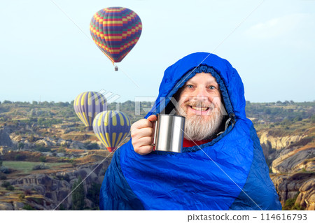 Happy bearded man in a sleeping bag with a cup of coffee against the backdrop of nature  114616793