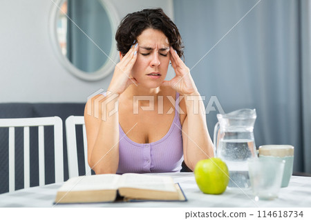 Young woman with headache sitting at table during reading book 114618734