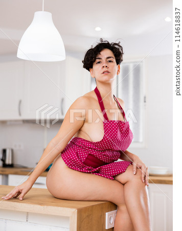 Flirting young female in red apron over naked body sitting on countertop in kitchen 114618837