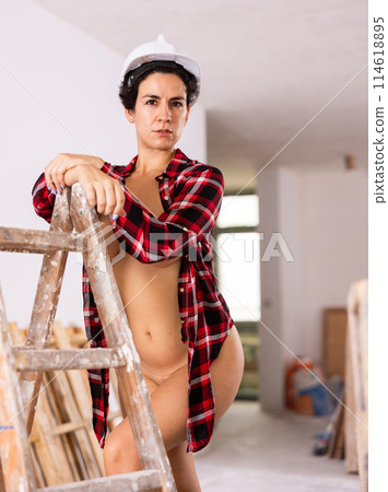 Attractive nudity builder in hardhat. Pretty girl wear unbuttoned red shirt sexy looking and leaning on stepladder 114618895