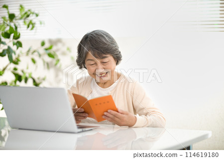 A senior woman using a computer while looking at her pension notebook, ending her life 114619180