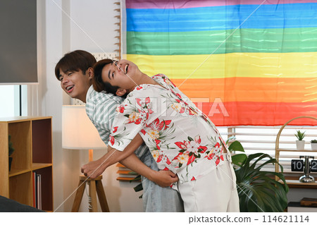 Playful gay couple giving piggyback ride for each other laugh joyfully in living room near rainbow LGBT Pride flag Playful gay couple giving piggyback ride for each other laugh joyfully in living room near rainbow LGBT Pride flag 114621114