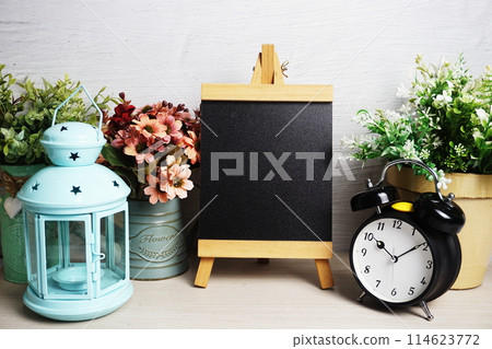 Easel mockup with blue landtern, alarm clock and flowers bouquet decoration on wooden background 114623772