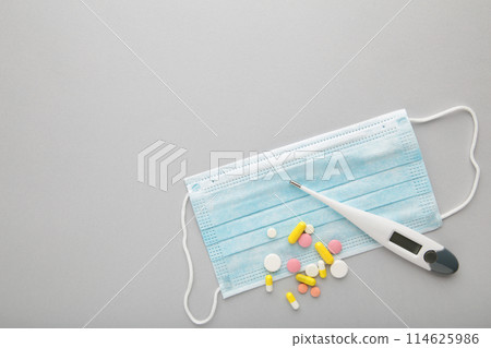 Medical thermometer and mask with pills on grey. Medical concept with copy space Medical thermometer and mask with pills on grey. Medical concept with copy space 114625986