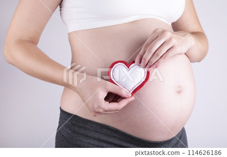 Pregnant women holding heart symbol placed on belly on grey background Pregnant women holding heart symbol placed on belly on grey background 114626186