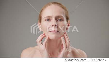 Real people - age, beauty, health and dry skin care concept - beautiful mature Caucasian middle aged woman in her 50s touching her face skin with a slight smile and good mood 114626447