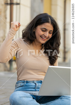 Indian happy woman working on laptop celebrate success win money sitting on urban street in city Indian happy woman working on laptop celebrate success win money sitting on urban street in city 114626885
